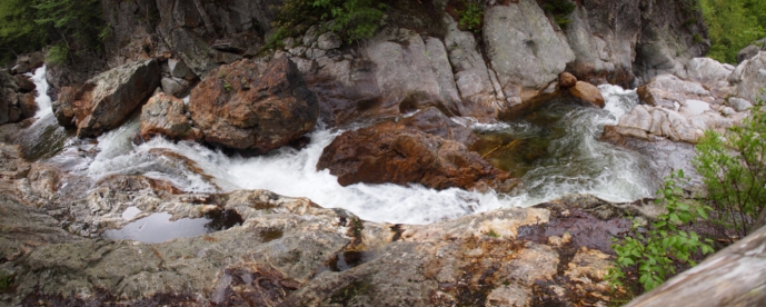 [Several photos stitched together showing a falls on the far left, then rushing water winding around rocks to a small pool and then the water flow disappearing into some large rocks on the right. Several large rust-colored boulders are on the far side against the hill while flatter rock is in the foreground.]